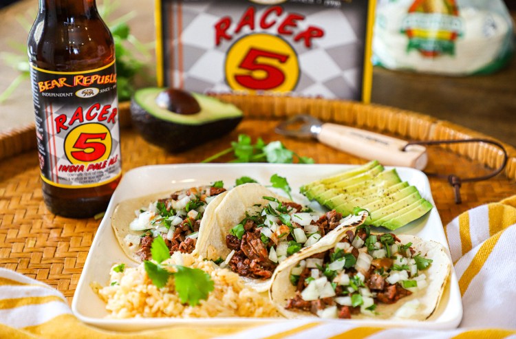 Bottle of Bear Republic Racer 5 IPA next to a plate of tacos, rice, and sliced avocado on a woven tray.